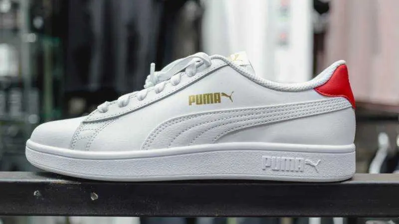 na zapatilla blanca de Puma, fotografiada de perfil, con el logotipo en dorado y un detalle rojo en el tal&oacute;n, sobre un fondo desenfocado de estilo comercial, UNSPLASH / ZAKARIA_ISSAAD